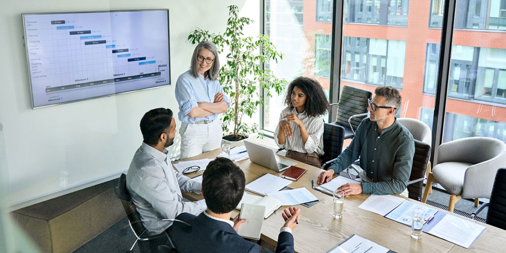 A diverse group of five professionals in a modern office meeting, gathered around a conference table with laptops, notebooks, and papers. A Gantt chart is displayed on a large screen as they discuss progress, while a woman stands leading the discussion. Large windows reveal an urban setting outside. Credit: Shutterstock