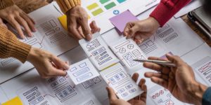 A team of people collaborates on a design project, reviewing hand-drawn wireframes and interface mockups. Several hands are visible, pointing and moving the sketches, which include layouts of various web or mobile app elements like icons, text boxes, and buttons. Color-coded sticky notes are also present on the table. Credit: Shutterstock