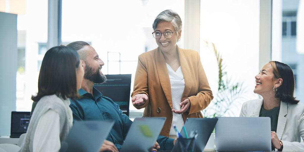 A group of four colleagues engaged in a lively discussion in a bright office. One woman stands, gesturing while speaking, as three seated coworkers listen attentively, smiling. Laptops are open on the table, and sunlight streams through large windows, creating a collaborative and positive atmosphere. Credit: Shutterstock