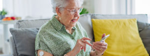 An elderly woman sits on a couch using her smartphone, smiling as she interacts with the device. She has a glucose monitor patch on her arm, indicating health monitoring. The setting is a cozy living room with a yellow pillow behind her. Image credit: Shutterstock.