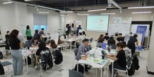 A collaborative workshop setting where several groups of people are engaged in discussions and activities around tables. Various tools like laptops, notebooks, and sticky notes are visible. A large screen in the background displays a coffee break time, and whiteboards and posters surround the room. The environment appears creative and interactive. Credit: Boyeun Lee