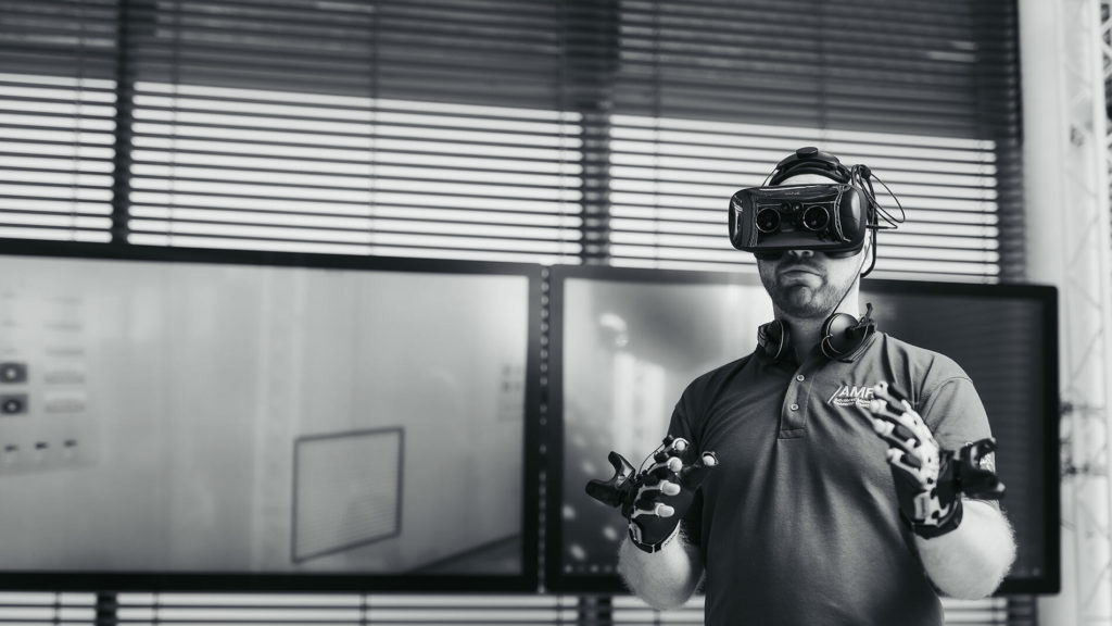 A man wearing a VR headset and data gloves stands in front of large display screens, engaging with virtual content in a high-tech environment. He wears an AMRC-branded polo shirt, indicating affiliation with the Advanced Manufacturing Research Centre. Credit: University of Sheffield, Advanced Manufacturing Research Centre (AMRC)