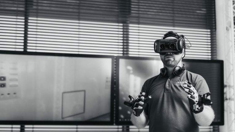 A man wearing a VR headset and data gloves stands in front of large display screens, engaging with virtual content in a high-tech environment. He wears an AMRC-branded polo shirt, indicating affiliation with the Advanced Manufacturing Research Centre. Credit: University of Sheffield, Advanced Manufacturing Research Centre (AMRC)