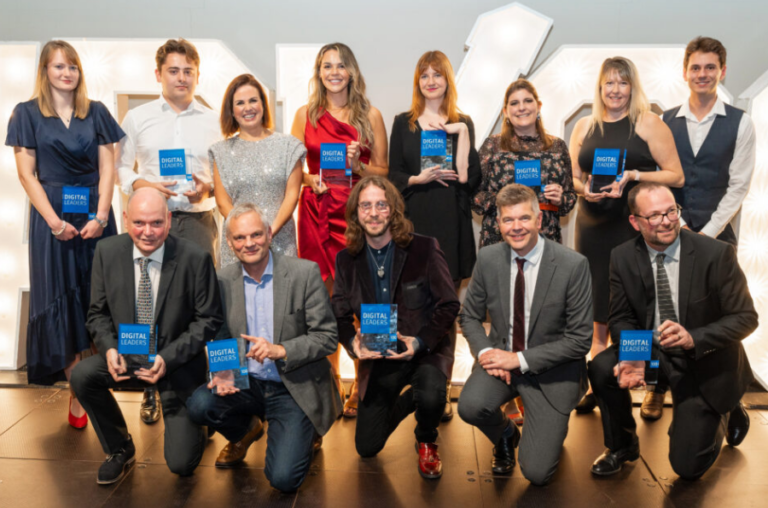 A group of eleven people posing on a stage, holding blue award plaques labeled “Digital Leaders.” Five women stand in the back row, and six men kneel in the front. Everyone is smiling, dressed in formal attire, under bright lights