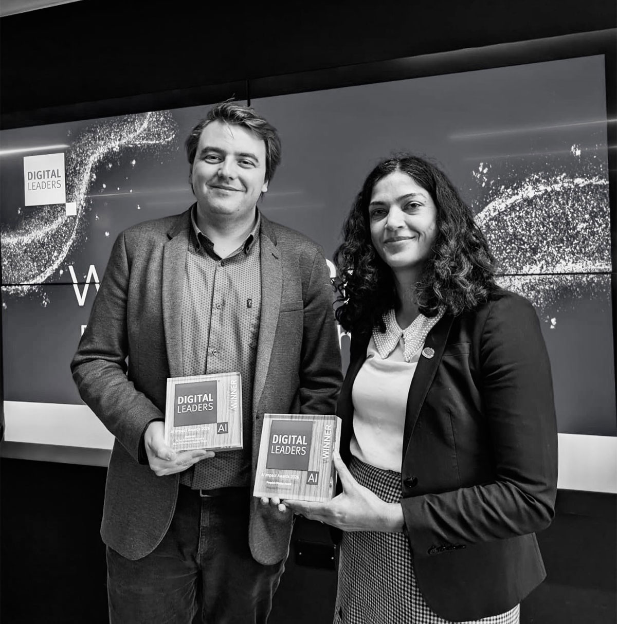 Two people standing side by side holding glass awards labelled Digital Leaders AI Winner, in front of a large Digital Leaders display screen with abstract light patterns, photographed at an awards event.